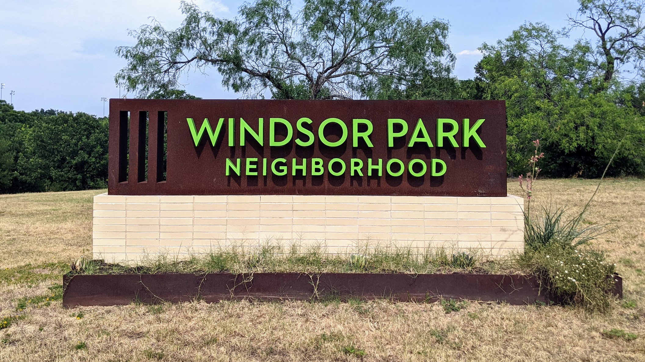 Long, rectangular rust brown sign with neon green letters reading 'Windsor Park Neighborhood'. There are 3 vertical lines on the left side of the sign.