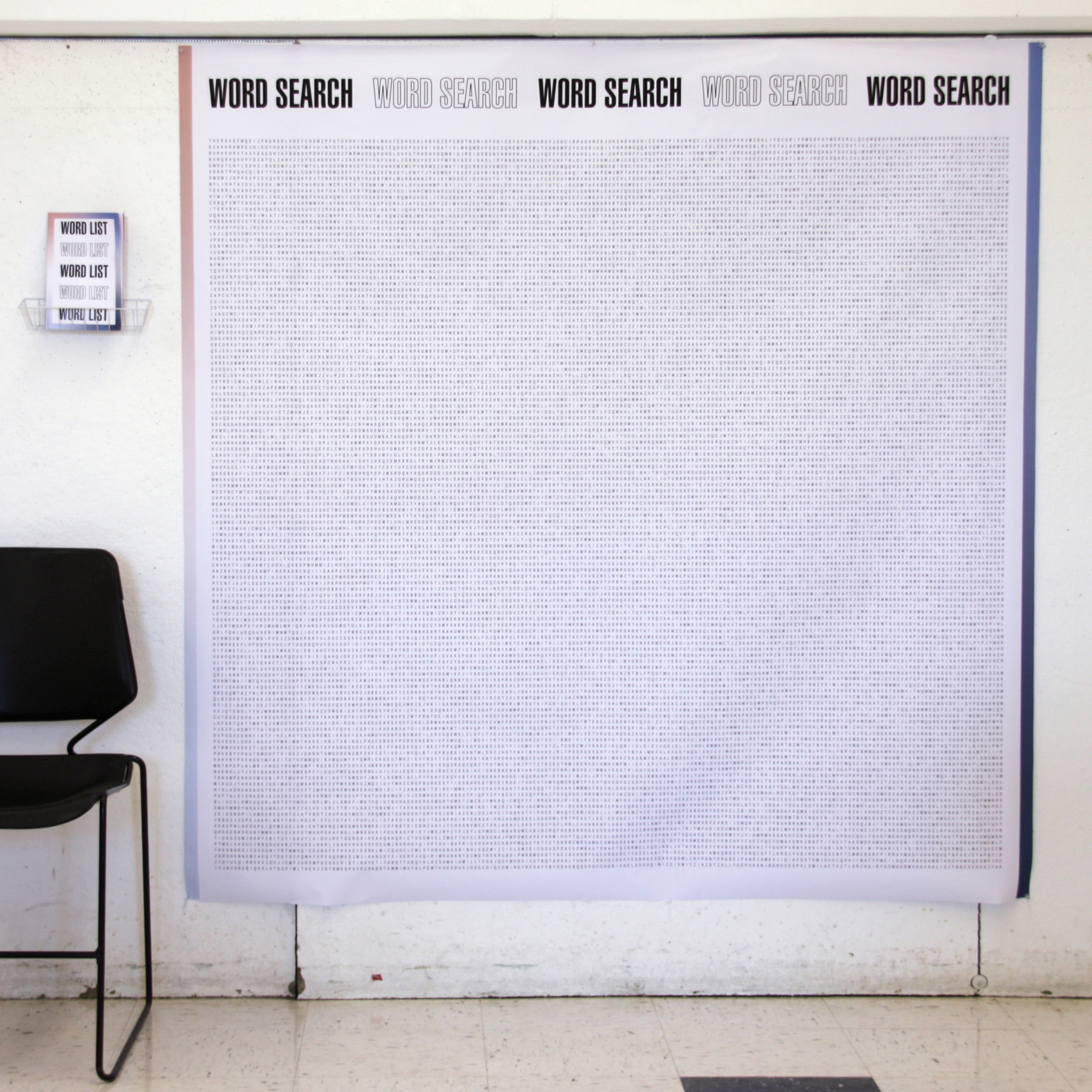 5 foot by 5 foot printed word search hangs on a wall. A chair sits next to it, in order to show scale. There is a small, wire basket pinned onto the wall next to the word search. In the basket is a printed booklet that reads 'Word List'.