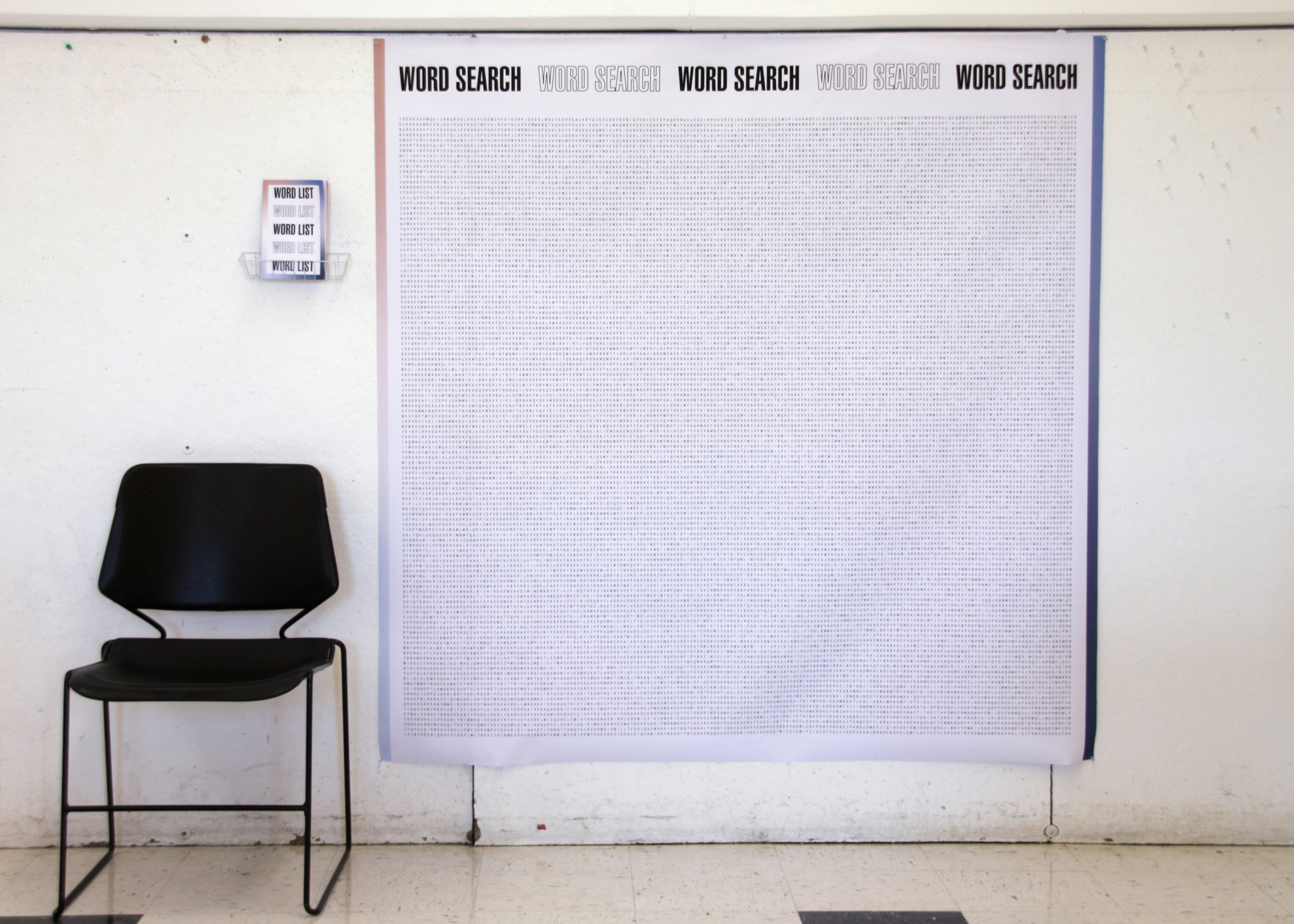 5 foot by 5 foot printed word search hangs on a wall. A chair sits next to it, in order to show scale. There is a small, wire basket pinned onto the wall next to the word search. In the basket is a printed booklet that reads 'Word List'.